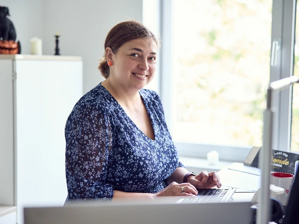 Eine lächelnde Frau mit blauer Bluse sitzt an einem Schreibtisch und tippt auf einem Laptop. Im Hintergrund sind ein Fenster und eine Reihe von dekorativen Gegenständen, darunter eine schwarze Katze und ein Kürbis, zu sehen.