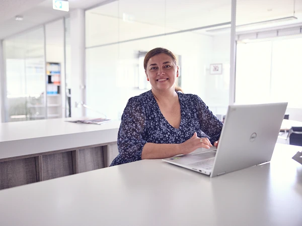 Eine Frau mit langen, braunen Haaren sitzt an einem Schreibtisch in einem modernen Büro und lächelt, während sie auf einen Laptop tippt. Hinter ihr sind Glaswände und Regale mit Akten zu sehen.