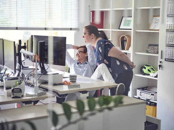 Zwei Personen in einem modernen Büro schauen auf einen Computerbildschirm, während eine der Personen auf etwas zeigt. Im Hintergrund sind mehrere Monitore und Büromaterialien sichtbar.