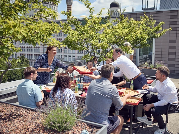Eine Gruppe von sieben Personen sitzt an einem Tisch auf einer Dachterrasse und genießt ein gemeinsames Essen. Eine Frau serviert Essen, während andere an dem Tisch lebhaft miteinander sprechen. Im Hintergrund sind Gebäude und Bäume zu sehen, und es herrscht sonniges Wetter.