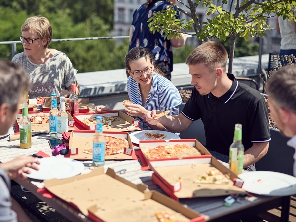 Eine Gruppe von fünf Personen sitzt an einem Tisch im Freien, umgeben von Grün, und genießt gemeinsam Pizza aus Pizzakartons, während sie sich unterhalten und lachen.