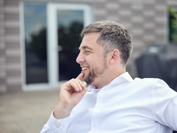 Ein Mann mit kurzen grauen Haaren trägt ein weißes Hemd und sitzt lächelnd auf einer Terrasse, während er nachdenklich seine Hand an sein Kinn legt. Im Hintergrund sind Fenster und eine unscharfe Grillstation sichtbar.