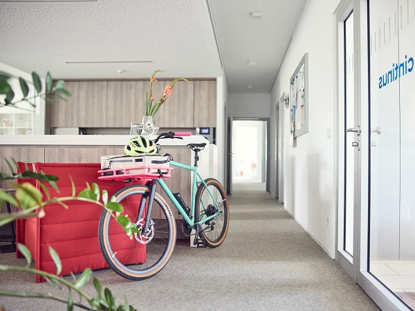 Ein Fahrrad mit einem Helm und einer Tasche steht in einem modernen Büroflur, in dem helle Wände und Teppiche zu sehen sind. Links ist ein rotes Sofa und eine Pflanze zu erkennen, während rechts eine Glastür mit dem Logo "citrinus" sichtbar ist.