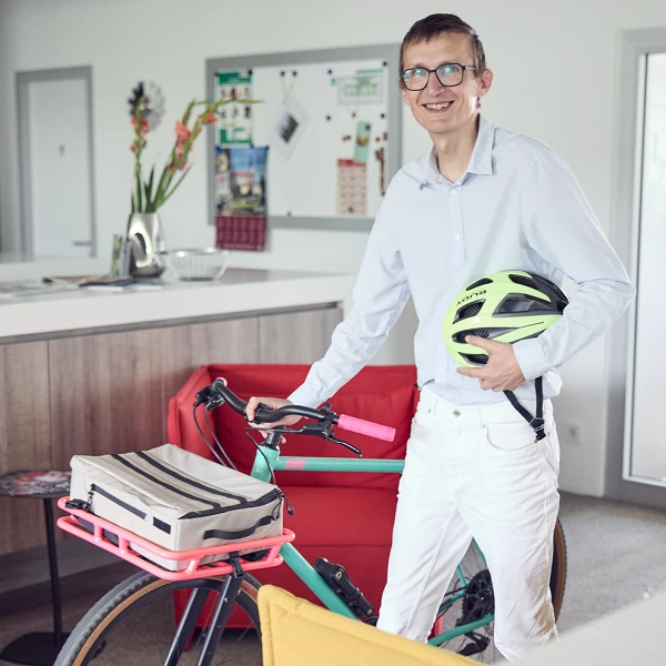 Ein Mann in einem hellen Hemd und mit Brille steht in einer modernen Küche und hält einen Fahrradhelm in der Hand. Neben ihm steht ein Fahrrad mit einem auffälligen Design. Im Hintergrund sind bunte Stühle und ein Tisch sichtbar, sowie einige Küchenutensilien.