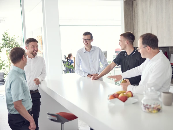 Eine Gruppe von fünf Männern steht um eine moderne Kücheninsel in einem hellen Büro, während sie lächeln und miteinander sprechen. Auf der Arbeitsfläche liegen frisches Obst und Süßigkeiten.